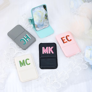 Pode incluir: Uma coleção de espelhos compactos personalizados em várias cores, incluindo cinza, preto, rosa e branco. Cada espelho apresenta iniciais únicas em cores contrastantes. Um espelho está aberto, mostrando uma imagem floral.