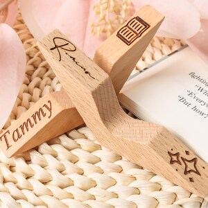 May include: Two wooden book page holders with engraved details. One has the name "Tammy", the other "Rinna". One has a book icon, and the other has star designs. The book holders are on a woven surface with a book.