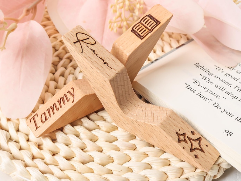 May include: Two wooden book page holders with engraved details. One has the name "Tammy", the other "Rinna". One has a book icon, and the other has star designs. The book holders are on a woven surface with a book.
