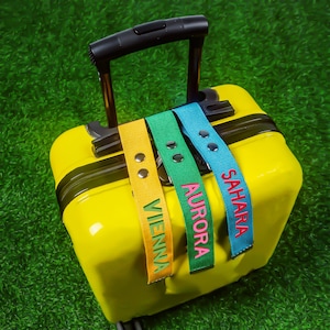 May include: A bright yellow suitcase with a black handle, adorned with three luggage tags. The tags are yellow, green, and blue, each displaying a name: "Vienna", "Aurora", and "Sahara". The names are embroidered in contrasting colours.
