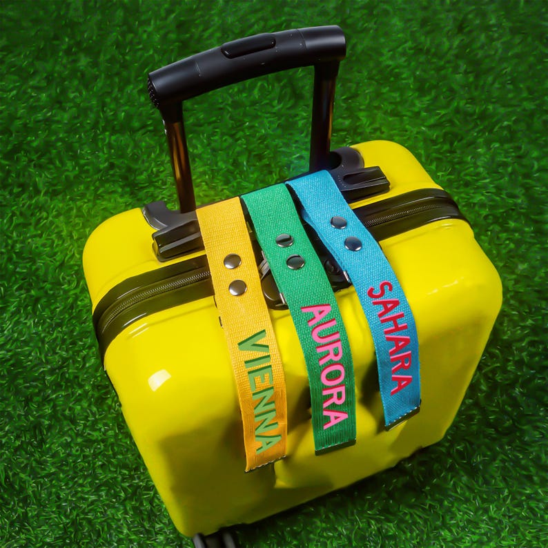May include: A bright yellow suitcase with a black handle, adorned with three luggage tags. The tags are yellow, green, and blue, each displaying a name: "Vienna", "Aurora", and "Sahara". The names are embroidered in contrasting colours.