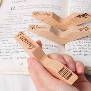 May include: Wooden book page holders with engraved names and designs. One holder is held over an open book. The holders are light brown and feature the names "Tammy", "Bailey", and "Raina". The "Tammy" holder has a book icon.