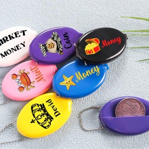 May include: A collection of colorful coin purses in various colors, each with a unique design and text. The purses include a white one with "Market Money", a purple one with a money bag, and a black one with a treasure chest.