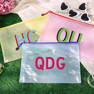 May include: Four mesh zipper pouches in yellow, pink, and blue, each with a different set of large, colorful letters. The pouches are on a green surface, with perfume bottles, makeup brushes, and heart-shaped sunglasses nearby.