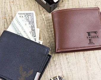 May include: Two leather wallets, one black and one brown, are displayed on a wooden surface. The black wallet has a silver corner accent and a design. The brown wallet is embossed with the name "FABRICE". A lighter and a pack of cigarettes are also visible.