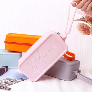 May include: A collection of rectangular pencil cases in various colors. The primary case is light pink with the name "Marcia" embossed on the front. Other cases are orange, gray, and light blue. Each case has a zipper closure and a wrist strap.