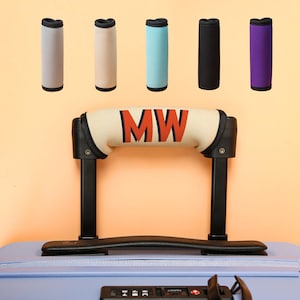 May include: A light blue suitcase with a black handle and a cream-colored handle wrap featuring the letters "MW" in orange and blue. Above, are five handle wraps in gray, beige, light blue, black, and purple.
