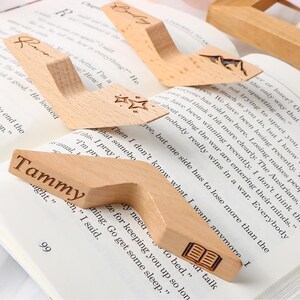May include: Wooden book holders with personalized names and star or book designs. These light-colored, angled book stands are placed on open books, ideal for readers. One holder has the name "Tammy" engraved.