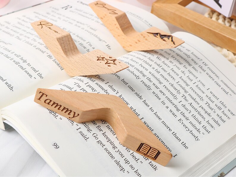 May include: Wooden book holders with personalized names and star or book designs. These light-colored, angled book stands are placed on open books, ideal for readers. One holder has the name "Tammy" engraved.