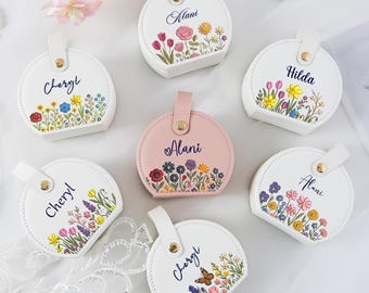 May include: A collection of personalized, round, white and pink leather cases. Each case features a floral design and a name in cursive. The cases have a handle and a gold snap closure.