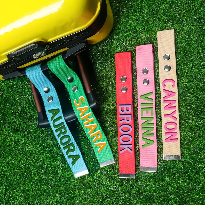 May include: A selection of brightly coloured luggage tags, each with a different name printed on it. The tags are blue, green, red, pink, and beige. The names read "Aurora", "Sahara", "Brook", "Vienna", and "Canyon". A yellow suitcase is in the background.