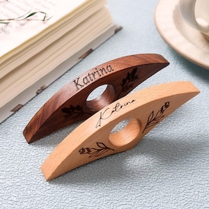 May include: Two wooden book page holders, one dark brown and one light wood. Each has a finger hole and the name "Katrina" engraved. The dark brown holder has a leaf design, and the light wood holder has a floral design.