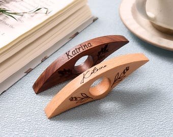 May include: Two wooden book page holders, one dark brown and one light wood. Each has a finger hole and the name "Katrina" engraved. The dark brown holder has a leaf design, and the light wood holder has a floral design.