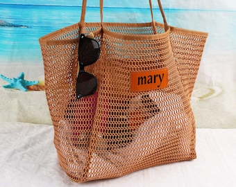 Op de afbeelding: Een bruine mesh tote bag met een leren patch waarop "mary" staat. De tas is gevuld met spullen, waaronder een zonnebril.