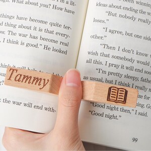 May include: A wooden bookmark with the name "Tammy" engraved on it. The bookmark also features a small, engraved image of an open book. The bookmark is rectangular and is held between the pages of a book.