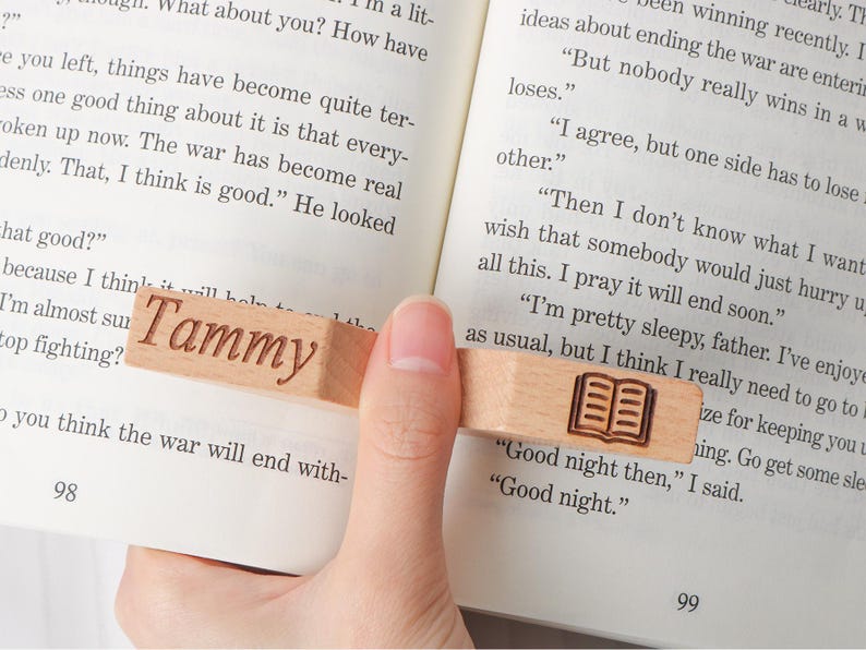 May include: A wooden bookmark with the name "Tammy" engraved on it. The bookmark also features a small, engraved image of an open book. The bookmark is rectangular and is held between the pages of a book.