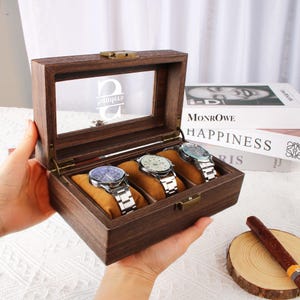 May include: A wooden watch box with a glass top and four slots for watches. The box has a personalized inscription on the glass top. The box is open and three watches are visible inside.
