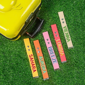 May include: A yellow suitcase sits on green grass, with five luggage tags in various colours. The tags, in yellow, orange, pink, and red, are labelled with the names "Sahara", "Aurora", "Breeze", "Lumina", and "Vienna".