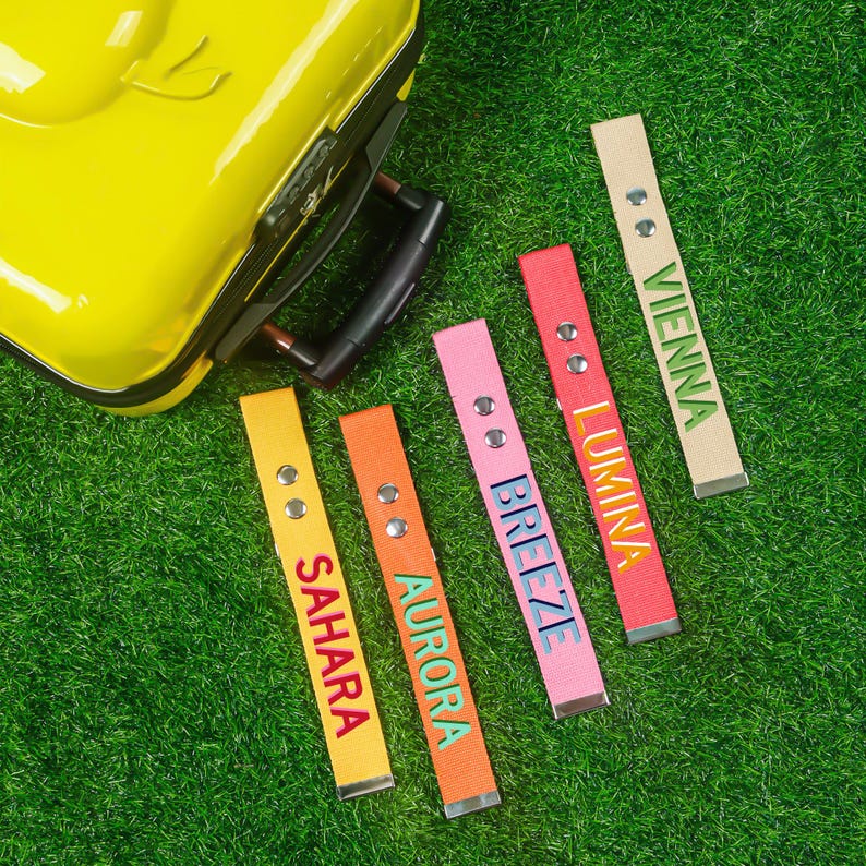 May include: A yellow suitcase sits on green grass, with five luggage tags in various colours. The tags, in yellow, orange, pink, and red, are labelled with the names "Sahara", "Aurora", "Breeze", "Lumina", and "Vienna".