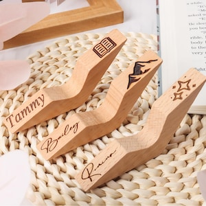 May include: Three wooden book page holders with engraved designs and names. One has a book icon, another a mountain climber, and the last has stars. The names "Tammy", "Bailey", and "Raina" are engraved on each holder.