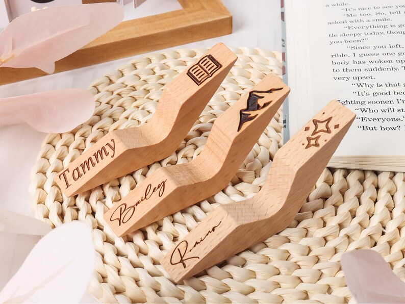 May include: Three wooden book page holders with engraved designs and names. One has a book icon, another a mountain climber, and the last has stars. The names "Tammy", "Bailey", and "Raina" are engraved on each holder.