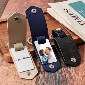 Peut inclure: Trois porte-clés en cuir beige, bleu marine et noir. Le porte-clés beige porte le texte "Your Photo" sur un insert blanc. Le porte-clés bleu marine affiche une photo d'un père et d'un enfant. Le porte-clés noir est gravé du nom "Noah".