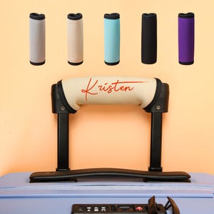 May include: A selection of luggage handle wraps in shades of beige, light blue, black, and purple. The main handle wrap is beige with the name "Kristen" in red script. These wraps are designed to fit over a suitcase handle for comfort and personalization.