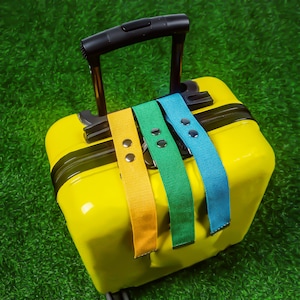 May include: A bright yellow suitcase with a black handle, featuring three straps in yellow, green, and blue. The straps are fastened with silver rivets. The suitcase is on a green surface.