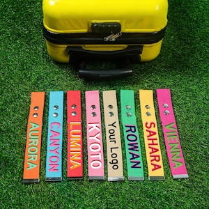 Puede incluir: Una maleta amarilla descansa sobre hierba verde, con una fila de etiquetas de equipaje coloridas debajo. Las etiquetas son naranjas, azules, rojas, rosas, beige, verdes, amarillas y fucsias, cada una con un nombre diferente.