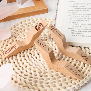 May include: Wooden book holders with personalized text, including "Tammy" and "Your Logo", are displayed on a woven mat. One holder has a book icon, another has stars. A book is open in the background.