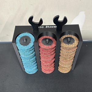 May include: A black plastic storage rack with the text Loc Stora printed on the front. The rack features three vertical cylindrical compartments holding stacks of blue, red, and tan abrasive sanding discs, topped with two black wrench icons.