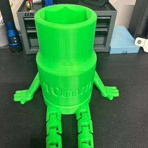 May include: A bright green 3D printed figure shaped like a socket tool with the text 10mm embossed on the front. The figure features a cylindrical body with a hexagonal opening at the top, two articulated arms extending to the sides, and two long jointed legs extending downward from the base.