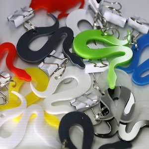 Pode incluir: Uma coleção de clipes de encadernação coloridos em forma de cobra em vermelho, amarelo, branco, verde, azul e cinza. Cada clipe é combinado com um clipe de encadernação de metal prateado. Os clipes estão espalhados em uma superfície branca.