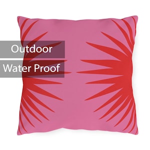 Puede incluir: Cojín rosa con un patrón geométrico rojo. El cojín está etiquetado como "Outdoor Water Proof".
