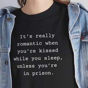 Puede incluir: Camiseta negra con el texto: "Es realmente romántico cuando te besan mientras duermes, a menos que estés en prisión." La camiseta se combina con una chaqueta vaquera y vaqueros azules.