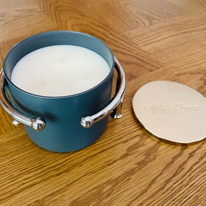 May include: A gray candle with a silver metal handle and a gold lid. The lid has the text "White Barn" embossed on it.