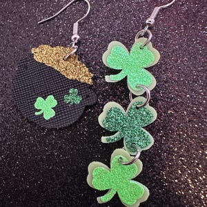 May include: St. Patrick's Day earrings. One earring features a black pot with a gold glitter rim and green shamrocks. The other earring has three layered green shamrocks with glitter accents, connected by silver rings and hooks.