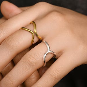 May include: Two minimalist rings, one gold and one silver, are shown on a hand. The gold ring has a curved, open design. The silver ring has a similar shape, creating a modern and elegant look. The rings are likely made of metal.