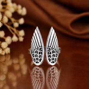 Puede incluir: Anillo plateado con diseño abierto que presenta dos formas de alas estilizadas. Cada ala tiene patrones de plumas detallados y una base curva. El anillo se encuentra sobre un fondo marrón borroso, con una superficie reflectante debajo.