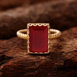 Puede incluir: Anillo dorado con una piedra preciosa rectangular de color rojo intenso. La piedra preciosa está enmarcada por un borde festoneado, y la banda tiene un diseño de cuerda trenzada. El anillo se muestra sobre una superficie de madera oscura.