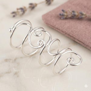 May include: A silver-colored ring with a unique, flowing design. The ring features multiple loops and swirls, creating an abstract and artistic look. The ring is displayed on a marble surface, with a small pink pouch and lavender sprigs in the background.