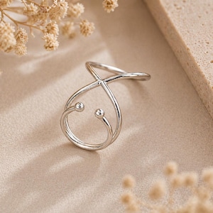 May include: A silver-colored ring with a unique, open design. The ring features a crisscross pattern with two small, round embellishments. The ring is displayed on a neutral-toned surface, with dried flowers in the background.