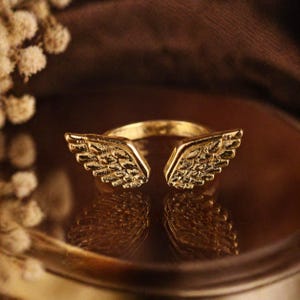 Puede incluir: Un anillo dorado con un diseño abierto, con dos formas de alas detalladas. Las alas tienen patrones texturizados en forma de plumas. El anillo se muestra sobre una superficie reflectante, resaltando el diseño de la joya.