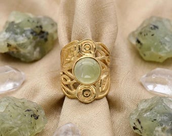 Può includere: Un anello color oro con una grande pietra rotonda verde pallido. L'anello ha un design ornato con motivi floreali e a spirale. L'anello è esposto su un panno beige, circondato da cristalli verdi e trasparenti.