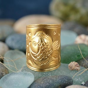 Könnte beinhalten: Ein goldfarbener, zylindrischer Ring mit einem geprägten Fischdesign. Der Ring befindet sich vor einem Hintergrund aus glatten, farbigen Steinen. Der Fisch ist mit Schuppen und Flossen detailliert, und der Ring hat eine polierte Oberfläche.