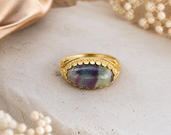 Puede incluir: Un anillo dorado con una piedra ovalada multicolor. La piedra presenta tonos de morado, verde y blanco. El anillo tiene una banda decorativa detallada con un borde festoneado y está sobre una superficie neutra.