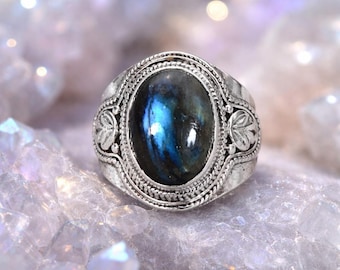 May include: A silver ring with an oval, iridescent blue gemstone. The ring has an ornate, detailed setting with leaf-like designs on the sides. The gemstone displays a play of blue and green colors.