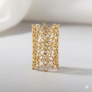 Peut inclure: Une bague dorée avec un motif floral. La bague est de forme cylindrique, avec des motifs de fleurs ajourées créant un aspect délicat et dentelé. La bague est posée sur un fond doux et neutre.