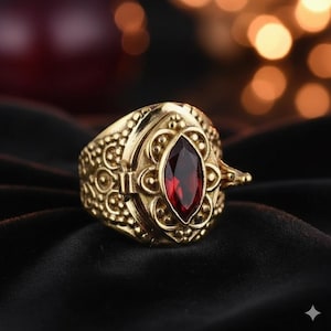 May include: A gold-colored ring with an ornate design and a large, oval, deep red gemstone. The ring has intricate detailing around the gemstone and on the band, with a clasp-like feature on the side. The ring is set against a dark background.