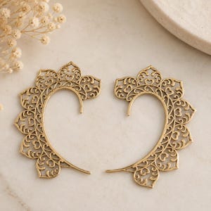 May include: A pair of gold-toned ear cuffs featuring an intricate filigree design with a scalloped edge. The metal pieces have a curved, open shape with detailed cutouts forming a repeating pattern throughout the surface of each individual ear cuff.